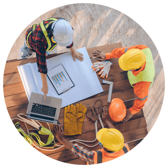Construction workers wearing hard hats are gathered around a wooden table to plan a project using printed documents and a laptop computer.