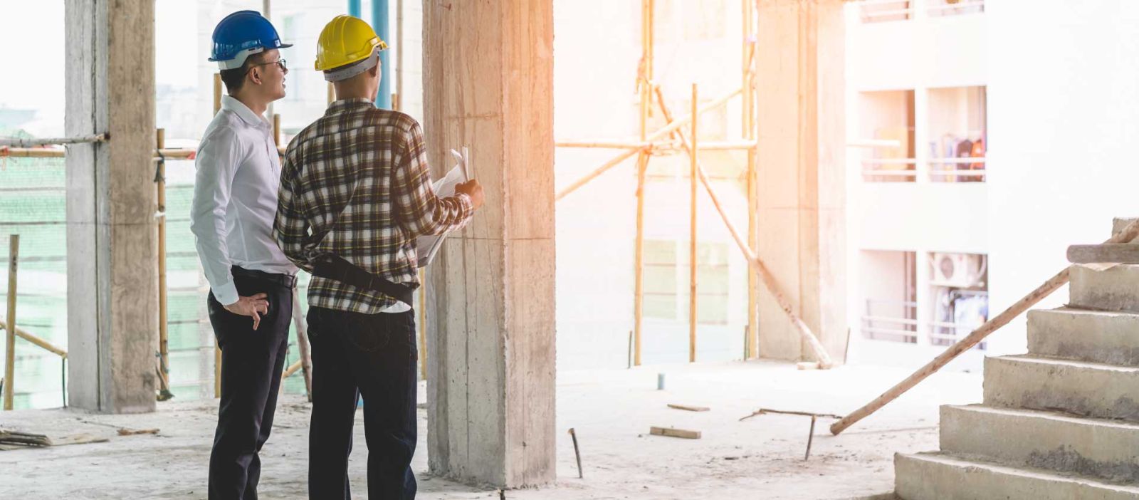 Two men wearing hard hats are looking off into the distance while standing in a construction site, one is holding architectural plans.